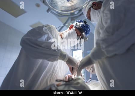 Dedicated surgeons perform intricate operation in modern hospital theater. Principal surgeon uses surgical loupes and headlamp, intently focused, with Stock Photo