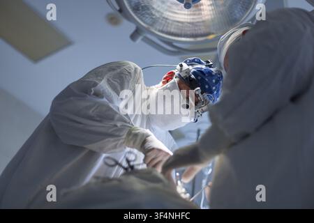 Focused medical team of surgeons performing intricate procedure in sterile hospital operating room under a bright lamp. Lead doctor wears surgical mag Stock Photo