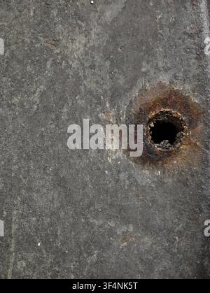 Close-up view of weathered concrete surface showing mottled gray texture, small white specks, and prominent rusted circular hole toward lower right, e Stock Photo