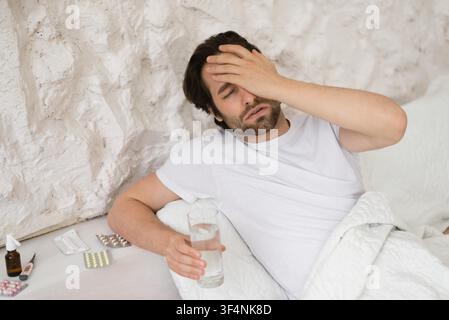 Sick Young Caucasian Man Sitting On Sofa Holding Glass Of Water, Covering Forehead, Healthcare Concept Stock Photo