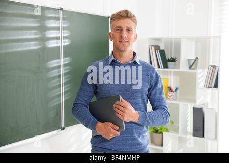 Portrait of smiling teacher with laptop near chalkboard in classroom Stock Photo