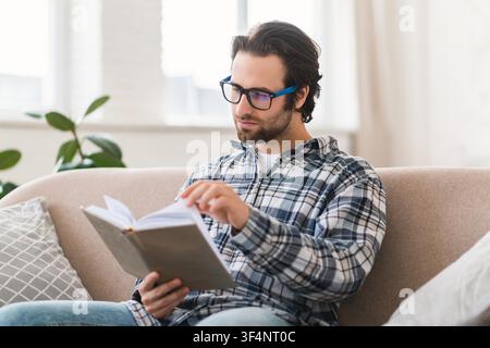 Focused Young Man Reading Book On Sofa At Home, Relaxing, Lifestyle Concept Stock Photo