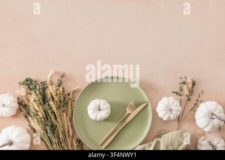 Elegant autumn table setting featuring white decorative pumpkin on ceramic plate with golden cutlery and dried grasses on soft beige background. Flat Stock Photo