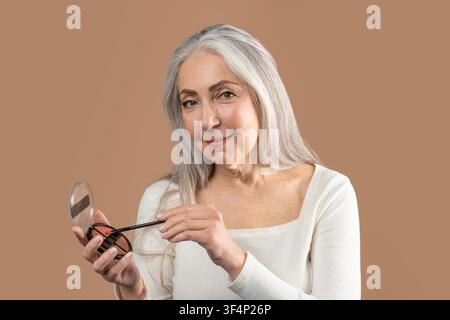 Senior Woman Using Makeup Mirror And Eyeliner, Smiling At Home, Skincare Concept Stock Photo