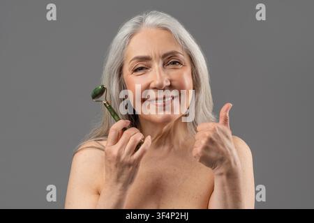 Happy Senior Woman Using Facial Roller And Showing Thumbs Up, Wellness Concept Stock Photo