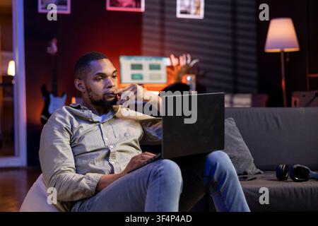 Black focused guy preparing to answer emails and finish daily tasks, enjoys working from the comfort of his modern apartment. Person navigating the internet on laptop, relaxing on the sofa. Stock Photo