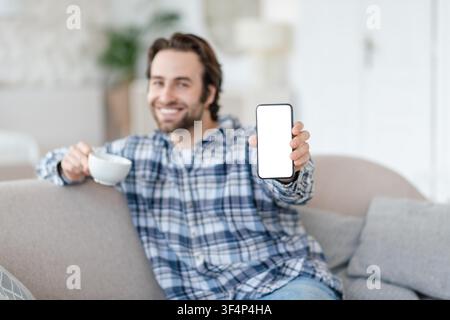 Smiling Caucasian Man Holding Smartphone With Empty Screen, Relaxing At Home Stock Photo