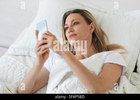 Young Woman Lying In Bed Using Smartphone, Relaxing At Home, Healthcare Concept Stock Photo