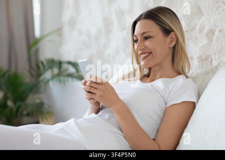 Smiling Young Woman Using Smartphone While Lying On Couch At Home, Lifestyle Concept Stock Photo