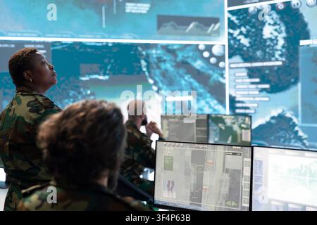 Command officer watches large surveillance wall from operations room. Military supervisor monitors intelligence feeds and enemy movement while operators work at computer stations Stock Photo