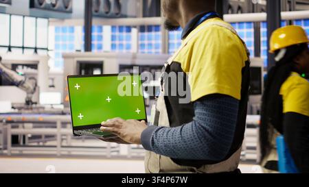 Engineer oversees solar panel production lines equipment using green screen laptop. Worker maintains photovoltaic assembly systems in automated industry 4.0 factory using chroma key notebook, camera A Stock Photo