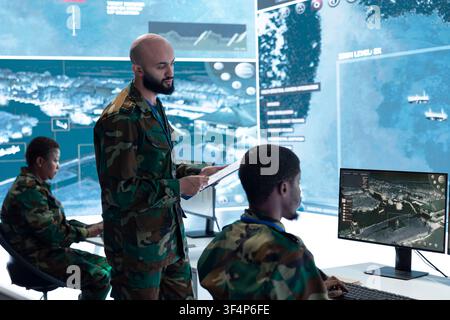 Military officer briefing personnel seated at command post workstation desks. Army squad team leader being provided mission information and intel data gathered by operators Stock Photo