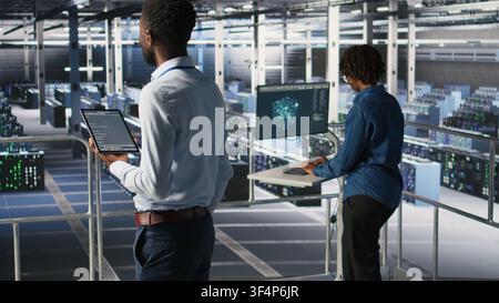 Data center programmer checking lines of code and colleague reviewing AI LLM visualizations. IT experts analyze neural networks outputs to ensure stable infrastructure operations, using tablet Stock Photo