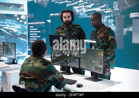 Military specialists present laptop data to seated surveillance operator in war room. Command post team colleagues compare intelligence information, analyzing tracked military targets Stock Photo