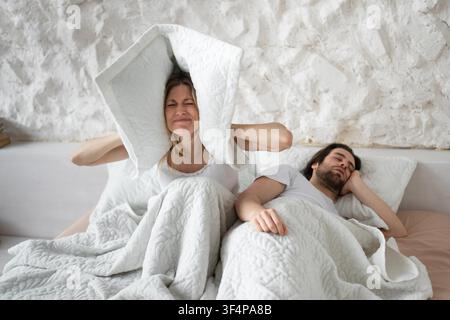 Stressed Young Woman Covering Ears With Pillow, Struggling To Sleep In Bed, Caucasian Male Partner Resting Stock Photo