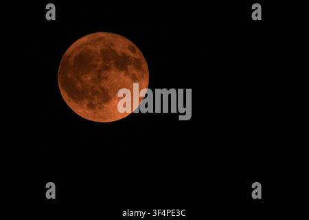 Detailed red buck full moon glowing against the dark night sky during July in Greece, showing clear surface craters and vibrant orange atmosphere. Stock Photo