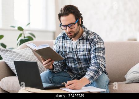 Focused Young Man Reading Book While Working On Laptop At Home, Study Concept Stock Photo