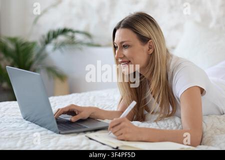 Smiling Young Woman Working On Laptop While Lying On Bed At Home, Remote Work Concept Stock Photo