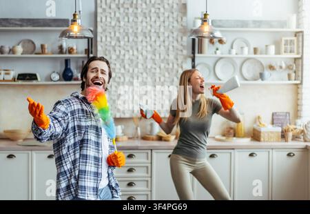 Happy Young Couple Cleaning Kitchen Together, Man Singing With Feather Duster, Woman Dancing Stock Photo
