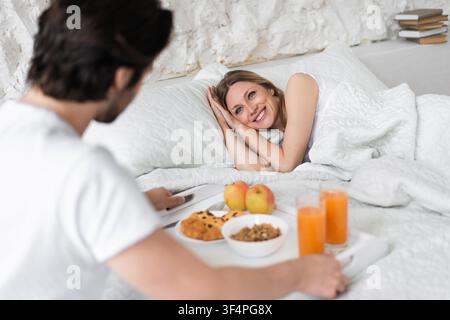 Happy Young Woman Smiling At Partner With Breakfast In Bed, Cozy Bedroom Setting Stock Photo