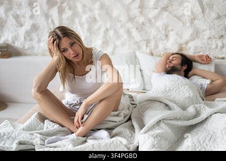 Worried Blonde Woman Sitting On Bed Next To Sleeping Man, Home, Relationship Conflict Concept Stock Photo