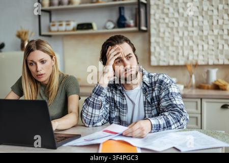 Worried Young Caucasian Couple Sitting At Kitchen Table With Laptop And Documents, Business Concept Stock Photo