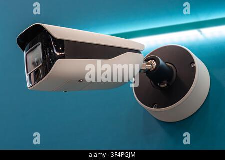 Modern white security camera with black lens cover, mounted on circular bracket, indoor showroom setting, vibrant blue background, clean professional Stock Photo