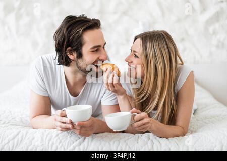 Happy Young Couple Smiling And Eating Breakfast In Bed, Morning Lifestyle Stock Photo