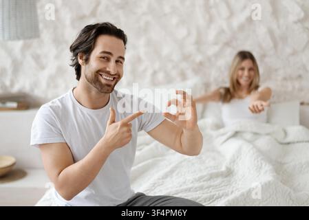 Happy Male And Female Couple Celebrating Together In Bedroom, Smiling And Pointing At Ring, Engagement Concept Stock Photo