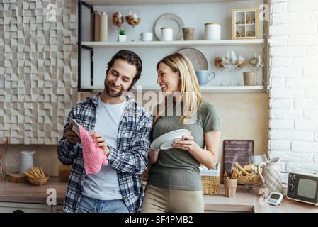 Happy Young Couple Smiling While Doing Dishes Together In Kitchen, Home Lifestyle Concept Stock Photo