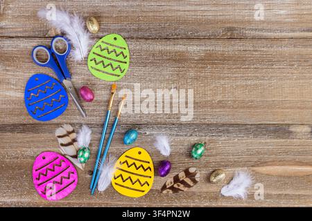 Vibrant Easter craft composition on the brown rustic wooden table with colored wooden eggs, paint brushes, scissors, candies, and feathers. Stock Photo