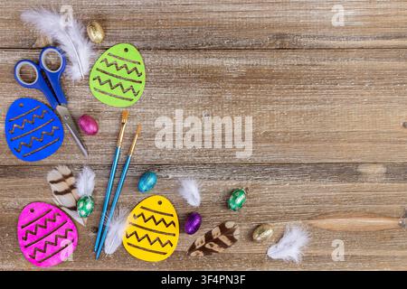 Vibrant Easter craft composition on the brown rustic wooden table with colored wooden eggs, paint brushes, scissors, candies, and feathers. Stock Photo