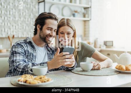 Happy Couple Enjoying Snacks And Coffee, Sitting At Kitchen Table, Using Smartphone Together Stock Photo