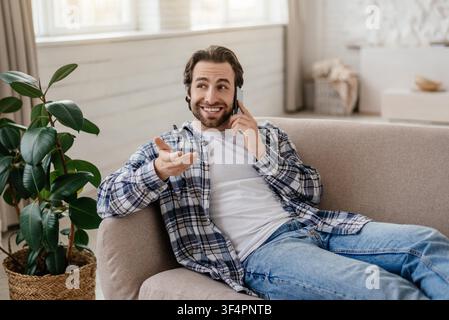 Happy Young Man Talking On Phone While Sitting On Couch In Modern Living Room Stock Photo