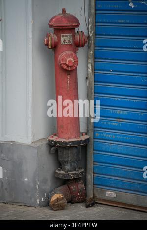 A weathered red fire hydrant stands vertically against a light gray concrete wall, to the left of a corrugated blue metal rolling shutter door. The hy Stock Photo