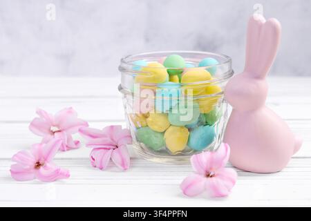 Easter decor on the white wooden table with ceramic pink bunny, colorful chocolate eggs in a glass jar and hyacinth pink flowers around. Stock Photo
