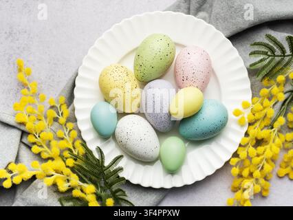 A selection of painted Easter eggs in pastel shades are elegantly displayed on a plate, accented with fresh yellow mimosa blooms. Stock Photo