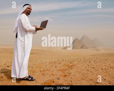 A confident Arab businessman in traditional dress smiles while using a laptop in the Egyptian desert with the Giza pyramids behind him. Authentic Middle East scene ideal for digital transformation - Photomontage Stock Photo