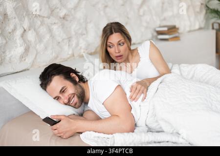 Happy Young Couple Sharing Moment In Bed, Smiling And Engaging With Smartphone, Cozy Setting Stock Photo