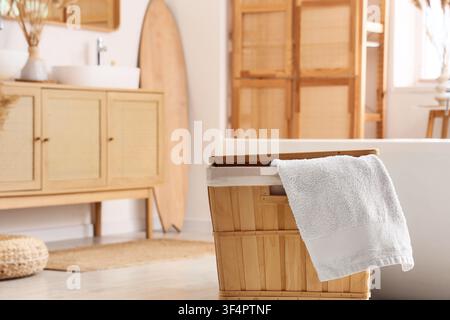White fluffy towel draped over a wooden laundry basket in a bright, modern bathroom with natural decor, the room features natural decor elements inclu Stock Photo
