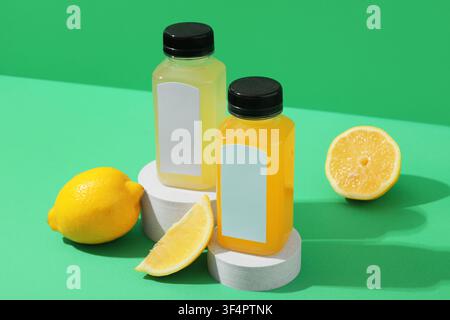 Two plastic bottles of fresh lemon and orange juice with blank labels on white pedestals with lemons on a green background Stock Photo