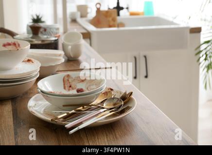 Pile of dirty dishes and cutlery on a wooden kitchen counter next to a sink, household chores and mess, the unwashed items are covered with food resid Stock Photo