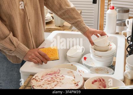 Person holding a yellow sponge and stacking dirty dishes in a kitchen sink, Household chores and cleaning Stock Photo