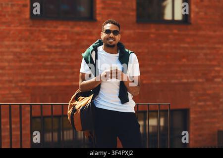 Smiling Young Black Man Using Smartphone With Backpack, Outdoor Urban Setting Stock Photo