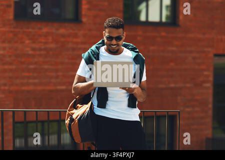 Happy Black Man Using Laptop Outdoors With Backpack, Urban Setting, Business Concept Stock Photo