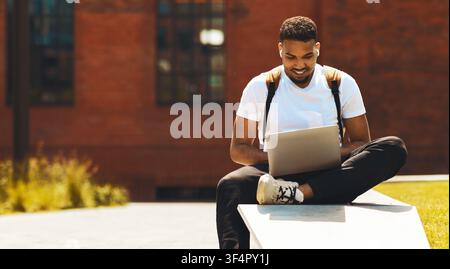 Smiling Black Man Working On Laptop Outdoors With Headphones, College Campus Setting Stock Photo