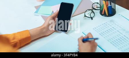 Unrecognizable Person With Smartphone At Desk, Writing Notes In Notepad, Office Setting Stock Photo
