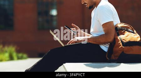 Young Man Using Laptop And Mobile Phone In Urban Setting, Focused Lifestyle Concept Stock Photo