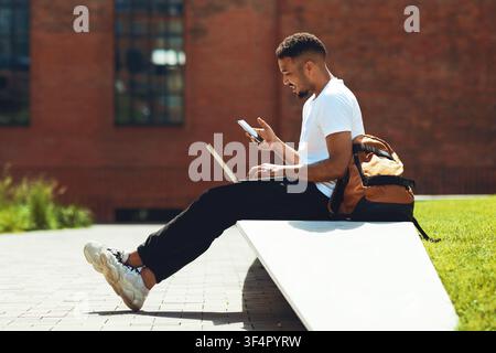 Young African American Man Using Laptop And Mobile Phone While Sitting Outdoors, Freelance Concept Stock Photo