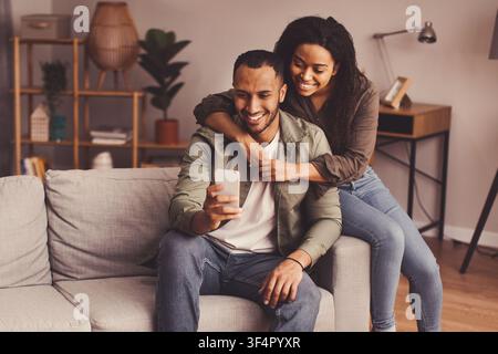 Happy Mixed Race Couple Smiling Together At Home, Engaging With Smartphone, Lifestyle Concept Stock Photo